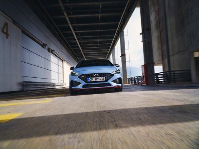 The new Hyundai i30 N from the front in Performance Blue colour driving along an industrial structure.